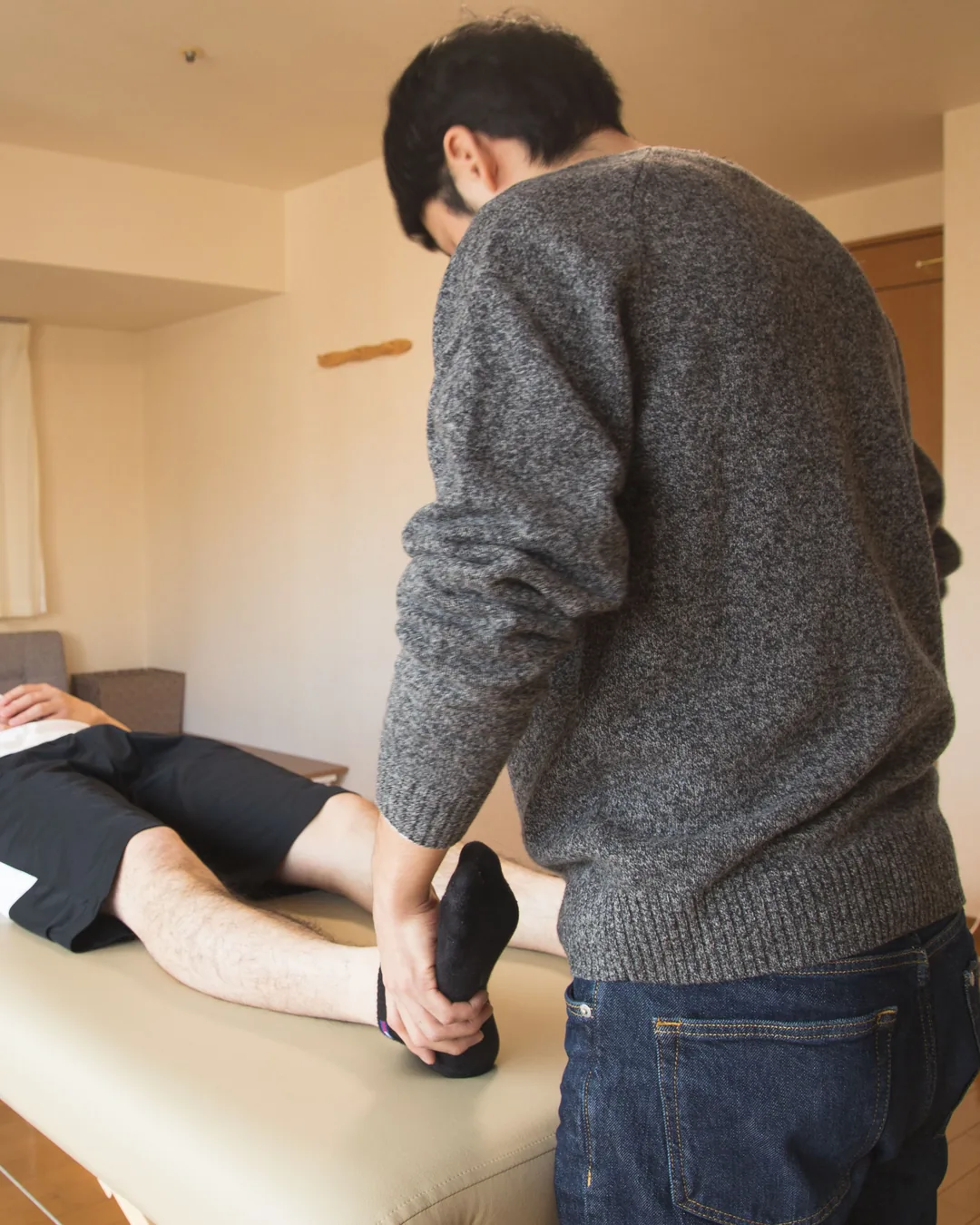 Un praticien mobilise la jambe d'une personne allongée sur une table de massage, dans une pièce à vivre